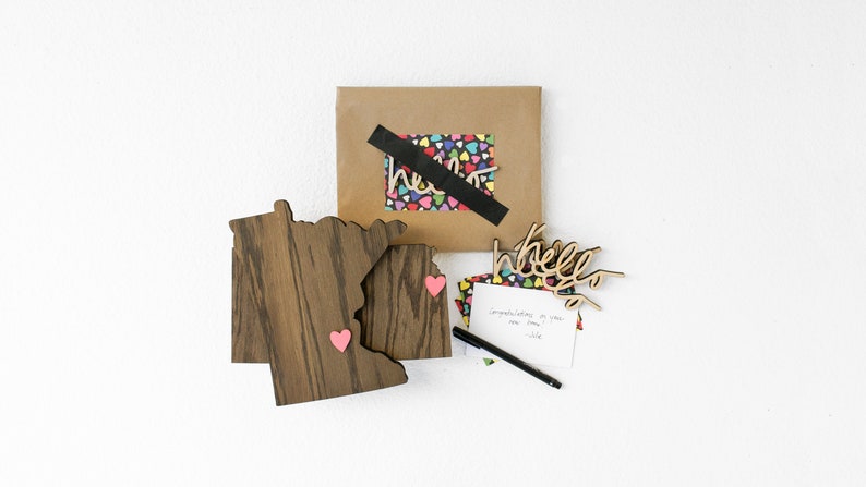May include: A brown wooden cutout of the state of Minnesota with two pink hearts attached. A brown paper envelope with a black tape seal and a colorful heart pattern inside. A white card with the word "hello" written on it. A black pen and a white card with a handwritten message.