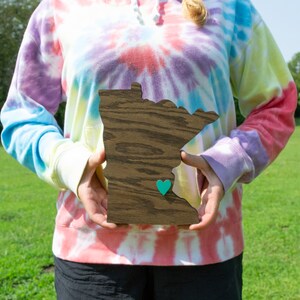 May include: A person is holding a wooden cutout of the state of Minnesota. The cutout is painted in a dark wood stain and has a small teal heart in the center. The person is wearing a tie-dye sweatshirt.