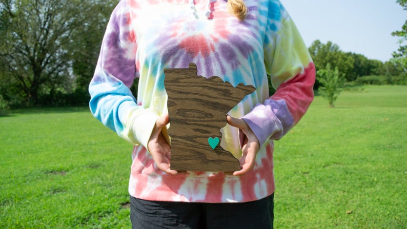 May include: A person is holding a wooden cutout of the state of Minnesota. The cutout is painted in a dark wood stain and has a small teal heart in the center. The person is wearing a tie-dye sweatshirt.