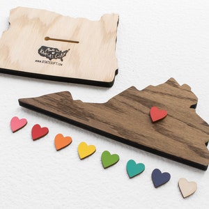 May include: Wooden cutout of the state of Virginia with a red heart placed on the state. A row of rainbow colored hearts are below the state. The back of the state cutout is made of dark wood. The text "Made in Kansas City" and "www.stategift.com" is printed on the back of the cutout.