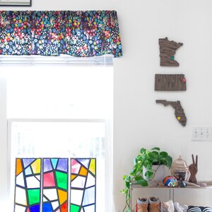 May include: A white wall with three wooden state outlines, Minnesota, Florida, and an unknown state, with colorful push pins. A stained glass window panel is on the left, and a plant is on a metal shelf on the right.
