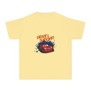 May include: A light yellow t-shirt featuring a red cartoon race car with a smiling face. The car is surrounded by blue and black accents, and the text "DRIVE & SURVIVE!" is above the car in orange.