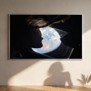 May include: Framed artwork depicting a silhouette of a person looking out a window at Earth from space. The Earth is visible through the window, showcasing blue oceans and swirling white clouds. The artwork is mounted on a beige wall.