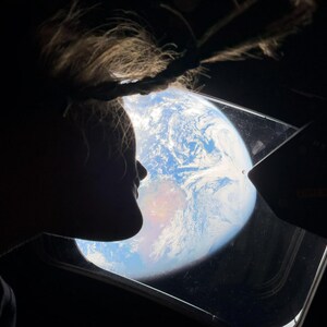May include: Silhouette of a person looking out a window at Earth from space. The Earth is a vibrant blue and white sphere with visible landmasses. The window frame and the person's hair are dark against the bright background.