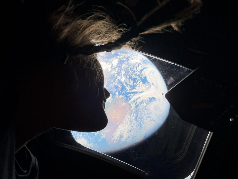 May include: Silhouette of a person looking out a window at Earth from space. The Earth is a vibrant blue and white sphere with visible landmasses. The window frame and the person's hair are dark against the bright background.