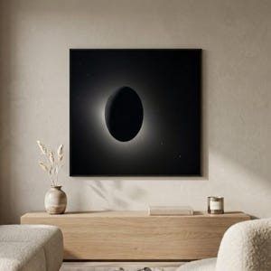 May include: A framed black and white print depicting a solar eclipse. The artwork features a dark oval shape surrounded by a bright halo against a black background. The print hangs above a wooden cabinet with decorative items.