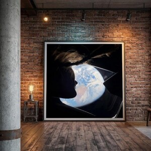 May include: A framed artwork featuring a silhouette of a person looking at Earth through a window. The image is set against a dark background, with the Earth visible in shades of blue and white. The room has a brick wall and wooden floor.
