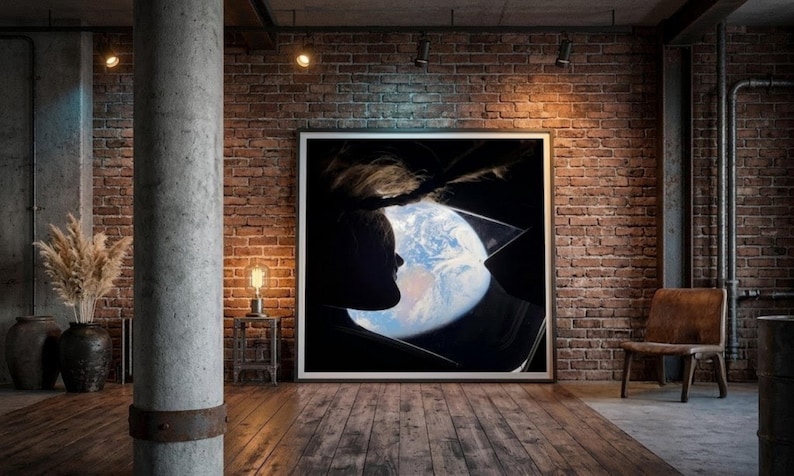 May include: A framed artwork featuring a silhouette of a person looking at Earth through a window. The image is set against a dark background, with the Earth visible in shades of blue and white. The room has a brick wall and wooden floor.