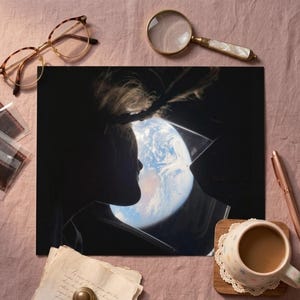 May include: A print of a person's silhouette looking at Earth from a window. The image is surrounded by a pair of glasses, a magnifying glass, a pen, a cup of coffee, film negatives, and a piece of paper.