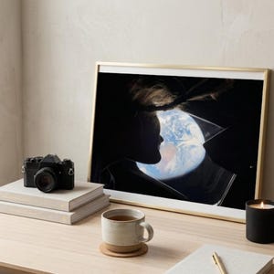 May include: A framed art print featuring a silhouette of a person with the Earth visible through a broken pane of glass. A black camera, books, a mug, a candle, and a plant sit on a wooden desk.