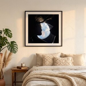 May include: A framed art print featuring a silhouette of a person's profile with the Earth visible within the head. The artwork is in a black frame and hangs above a bed with beige bedding and decorative pillows. A potted plant and side table are in the foreground.