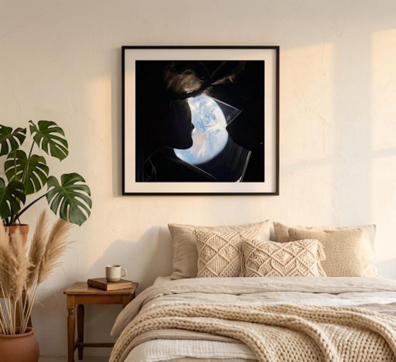 May include: A framed art print featuring a silhouette of a person's profile with the Earth visible within the head. The artwork is in a black frame and hangs above a bed with beige bedding and decorative pillows. A potted plant and side table are in the foreground.