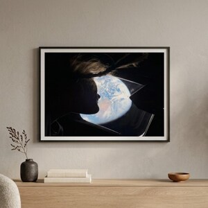 May include: Framed art print featuring a silhouette of a person looking at a depiction of Earth. The artwork is set against a dark background, with the planet's blue and white colors visible. The print is in a black frame, displayed on a light wood cabinet.