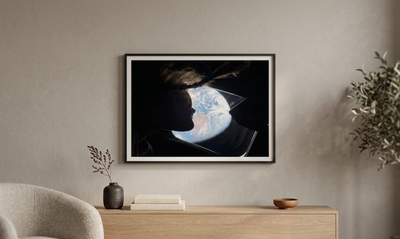 May include: Framed art print featuring a silhouette of a person looking at a depiction of Earth. The artwork is set against a dark background, with the planet's blue and white colors visible. The print is in a black frame, displayed on a light wood cabinet.
