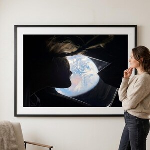 May include: A framed print featuring a silhouette of a person looking at Earth from space. The artwork is displayed on a white wall, with a woman standing nearby, gazing at the print. A neutral-toned armchair and an olive tree are also visible.