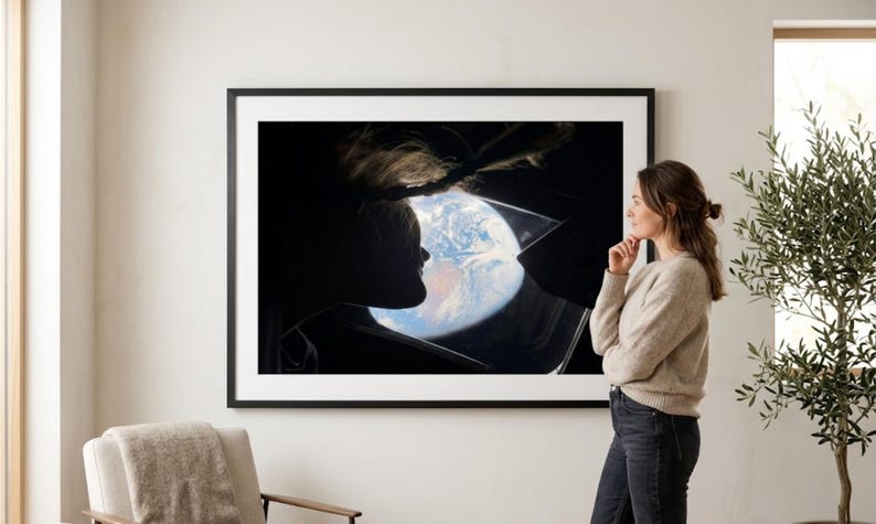 May include: A framed print featuring a silhouette of a person looking at Earth from space. The artwork is displayed on a white wall, with a woman standing nearby, gazing at the print. A neutral-toned armchair and an olive tree are also visible.