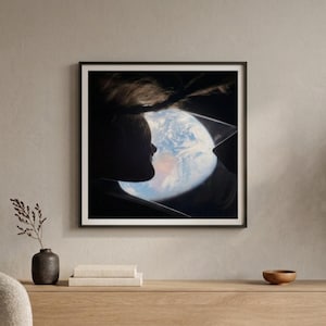 May include: A framed artwork depicts a silhouette of a person looking at Earth from a window. The Earth is visible with blue oceans and white clouds. The artwork is in a black frame, hanging on a neutral-colored wall.