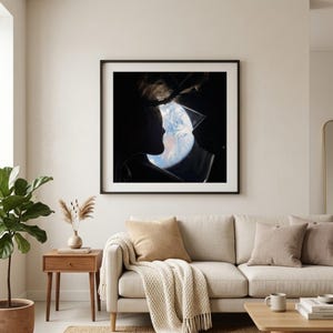 May include: A framed art print featuring a silhouette of two people kissing, with the Earth visible in the background. The artwork is displayed above a beige sofa with throw pillows and a blanket. A wooden side table with a plant and vase is to the left.