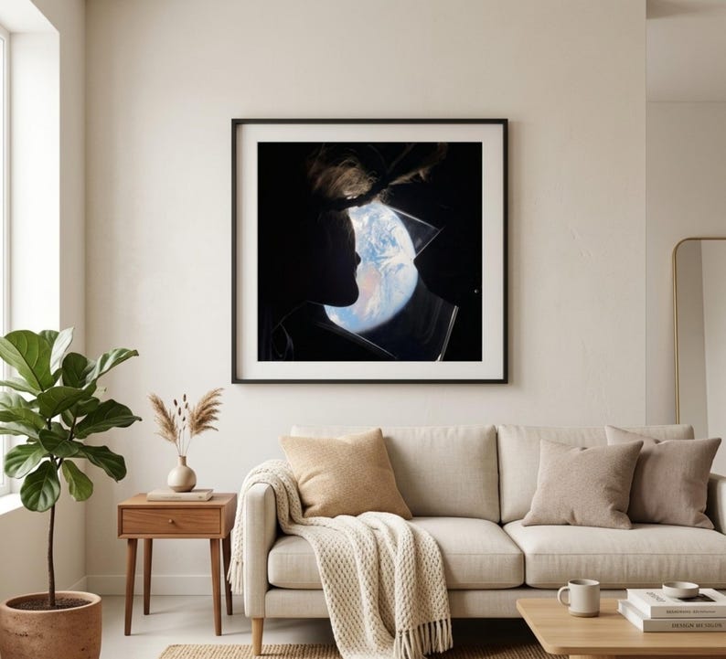 May include: A framed art print featuring a silhouette of two people kissing, with the Earth visible in the background. The artwork is displayed above a beige sofa with throw pillows and a blanket. A wooden side table with a plant and vase is to the left.