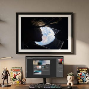 May include: A home office setup with a desk, computer, and framed artwork. Comic books and a desk lamp are visible. A figurine and a small potted plant add decorative touches. The computer screen displays a design program.