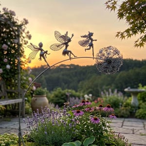 Könnte beinhalten: Eine Metallgarten-Skulptur mit drei Feenfiguren mit filigranen Flügeldesigns, die auf einer gebogenen Stange sitzen. Eine große, kugelförmige Löwenzahn-Skulptur ist ebenfalls Teil des Designs. Die Szene spielt in einem Garten mit Blumen und einem Sonnenuntergang.