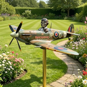 Puede incluir: Maqueta de avión con diseño de camuflaje, con el logo de Iron Maiden y una figura de piloto. El avión está montado sobre un soporte dorado, en un jardín con flores. La envergadura es de aproximadamente 100 cm.