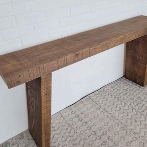 Peut inclure: Une longue console rectangulaire en bois avec une finition naturelle. La table a un dessus épais et deux pieds robustes, mettant en valeur le grain du bois. La table mesure environ 183 cm de long.