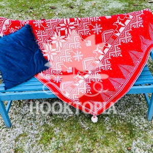 May include: A bright red knit blanket featuring a white geometric snowflake and house pattern, draped over a light blue wooden bench. A solid dark blue square pillow with small pom-pom trim sits on the blanket. The text HOOKTOPIA is visible.
