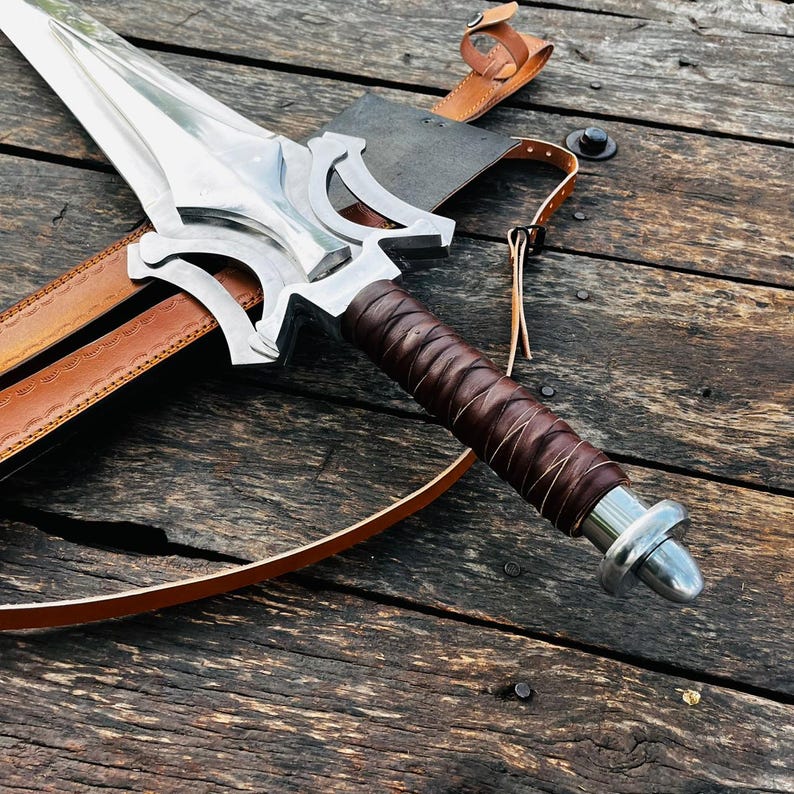 Handmade fantasy sword style knife with a long polished blade displayed on rustic wooden surface, showing the full product clearly along with premium leather sheath. This unique collector piece features a bold artistic guard design and a leather wrapped handle for a strong grip, making it an ideal gift for husband, anniversary present, or display item for collectors who appreciate detailed craftsmanship and standout design.