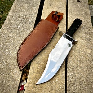 Può includere: Un grande coltello in acciaio lucido con manico nero e dettagli in ottone, accanto a un fodero in pelle marrone. La lama ha un bordo seghettato. Il fodero è impresso con un motivo decorativo e ha un passante per cintura.