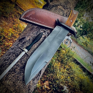 May include: A large, stainless steel knife with a wooden handle and a leather sheath. A sharpening steel with a black handle and a leather sheath are also present. The knife is resting on a tree trunk.