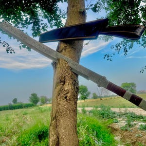 May include: A large, silver-colored sword with a dark red and silver handle is leaning against a tree. A black leather sheath is behind the sword. The sword has a long, rectangular blade with a textured surface. The background shows a green field and a blue sky.
