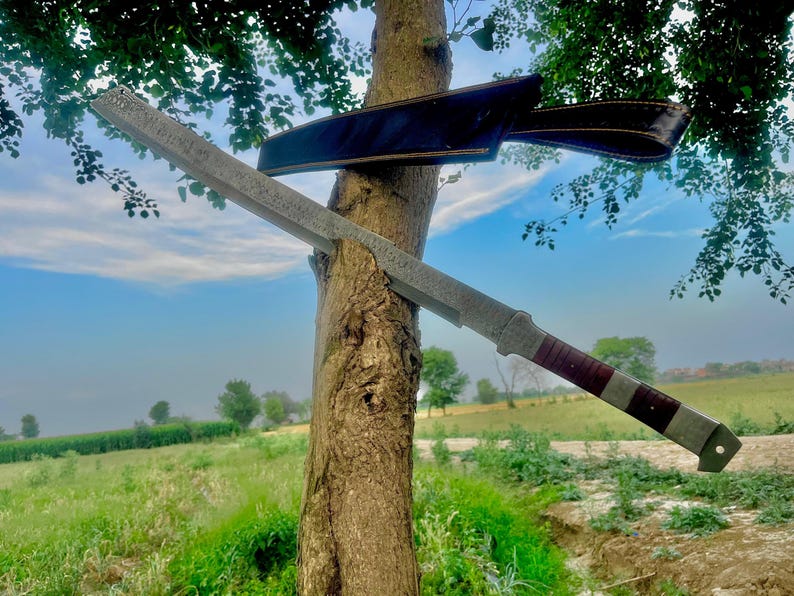 May include: A large, silver-colored sword with a dark red and silver handle is leaning against a tree. A black leather sheath is behind the sword. The sword has a long, rectangular blade with a textured surface. The background shows a green field and a blue sky.