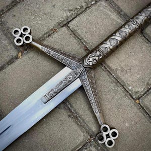 May include: A detailed, silver-colored broadsword with intricate engravings. The hilt features a lion emblem and decorative patterns. The crossguard and pommel have a unique, openwork design. The blade is long and polished, reflecting the light.