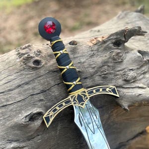 Könnte beinhalten: Ein dekoratives Schwert mit einem schwarzen Griff, der mit goldfarbenen Akzenten umwickelt ist. Der Knauf hat einen goldenen Parier mit schwarzen Details. Der Knauf ist schwarz mit einem roten Edelstein. Die Klinge hat eine silberne Oberfläche.