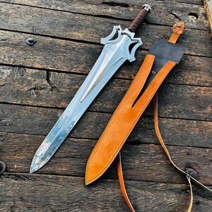 Custom handmade fantasy dagger style knife paired with a high quality leather sheath, captured in a clean top view to highlight overall craftsmanship. The long blade has a sleek reflective finish while the handle is wrapped in leather for comfort and control. This piece is perfect for collectors, display setups, or as a memorable gift for husband, boyfriend, or special occasions.