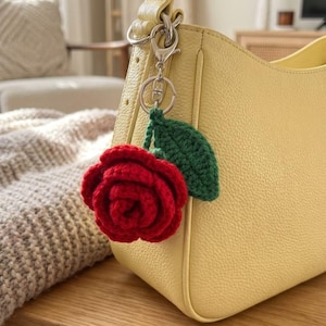 May include: A handmade red crochet rose with a green leaf attached to a silver keychain. The rose is hanging from a yellow leather handbag. The background includes a knitted blanket and a chair.