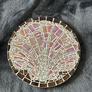 May include: A round mosaic coaster featuring a seashell design. The mosaic is made of iridescent pink, purple, and white tiles, framed by a brown border. The coaster is on a gray surface.