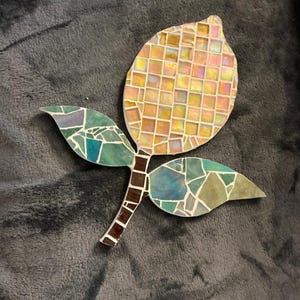 Puede incluir: Una obra de arte en mosaico con forma de flor estilizada. El cabezal de la flor es una mezcla de baldosas cuadradas amarillas, naranjas y rosas. El tallo es marrón y las hojas son piezas de mosaico verdes y azules. La obra de arte está sobre un fondo gris.