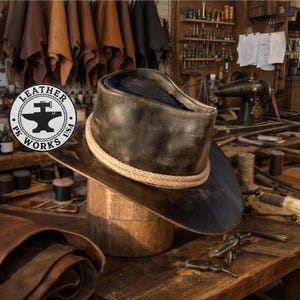 Pode incluir: Um chapéu de cowboy de couro castanho escuro com uma faixa de corda bege claro está em um bloco de chapéu de madeira. O chapéu tem uma aba larga e uma copa alta. A imagem inclui o logotipo "Leather Works USA" e uma oficina.