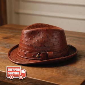 May include: A brown fedora hat featuring a textured ostrich-skin pattern. The hat includes a matching brown leather band around the crown, secured with a small, metallic gold-toned buckle. The surface of the hat displays a distinct quill-dot texture.