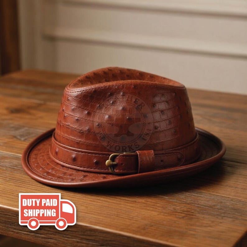 May include: A brown fedora hat featuring a textured ostrich-skin pattern. The hat includes a matching brown leather band around the crown, secured with a small, metallic gold-toned buckle. The surface of the hat displays a distinct quill-dot texture.