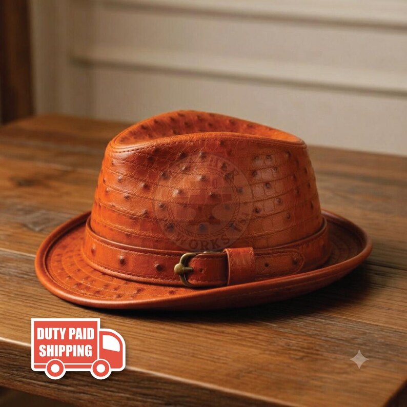 May include: A cognac-colored fedora hat crafted from textured leather with a distinct quill pattern. The hat features a matching leather band around the crown, secured by a small, metallic buckle on the side, creating a structured silhouette.