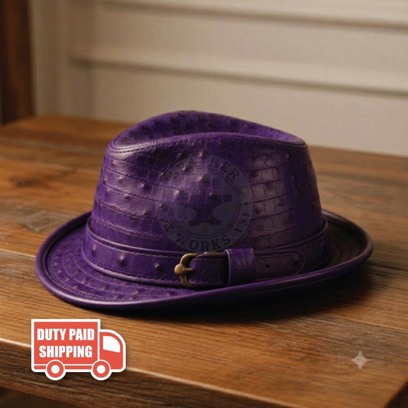 May include: A vibrant purple fedora hat featuring a textured ostrich-skin pattern. The hat includes a matching purple leather band around the crown, secured with a metallic brass-toned buckle. A circular logo stamp is visible on the front crown.