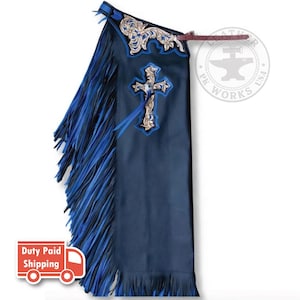 May include: Black leather chaps featuring long blue and black fringe along the side. The upper section and a central cross emblem display intricate tan floral tooling with blue accents. A brown leather strap is attached at the top waist area.