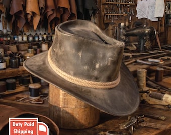 May include: A distressed dark gray leather hat with a wide brim and a rounded crown, featuring a thick, light tan braided rope band wrapped around the base of the crown. The hat has a weathered, worn-in appearance with visible texture and shading.