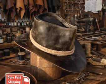 May include: A dark brown, distressed leather hat with a wide brim and a thick, light tan braided rope band encircling the crown. The hat features a weathered, textured finish and a structured, cylindrical crown shape with a slightly curved brim.