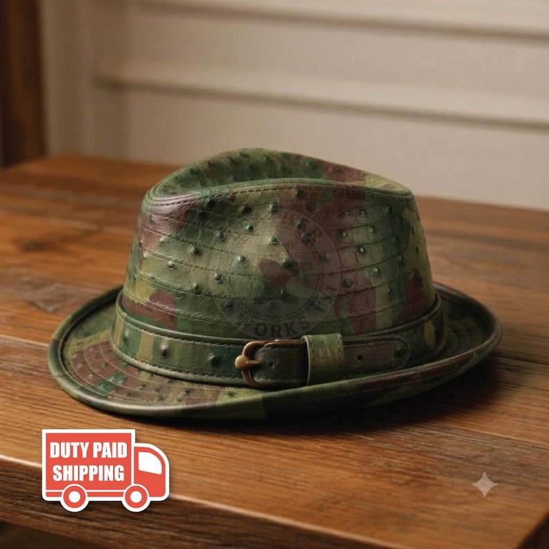 May include: A fedora hat featuring a green and brown camouflage pattern with a textured, quill-like surface finish. The hat includes a matching leather-style band around the base, secured with a small, dark metal buckle on the side.
