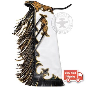 Pode incluir: Chaps de couro branco com acabamento em couro preto e detalhes florais dourados. O painel lateral apresenta longas franjas pretas e douradas. Uma tira de cinto de couro preto está presa no topo. Um logotipo circular diz LEATHER PK WORKS USA.
