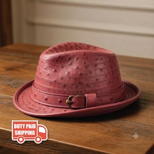 May include: A dusty rose fedora hat featuring a textured ostrich skin pattern. The hat includes a matching band around the crown with a metallic buckle detail. The surface of the hat displays a repeating quill follicle pattern across the entire exterior.